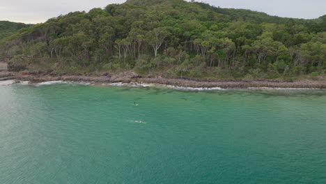 Szenische-Luftdrohne-Eines-Sportlers,-Der-Vor-Der-Küste-Mit-Einem-Stand-up-Paddle-Boarder-Im-Meer-Schwimmt,-4k-Noosa,-Australien,-Zeitlupe