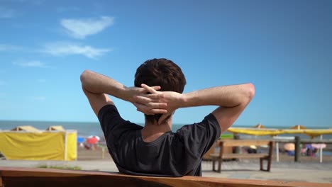 rear of a man with hands over back of his head sitting on wooden bench at summer beachfront of monte hermoso, buenos aires, argentina