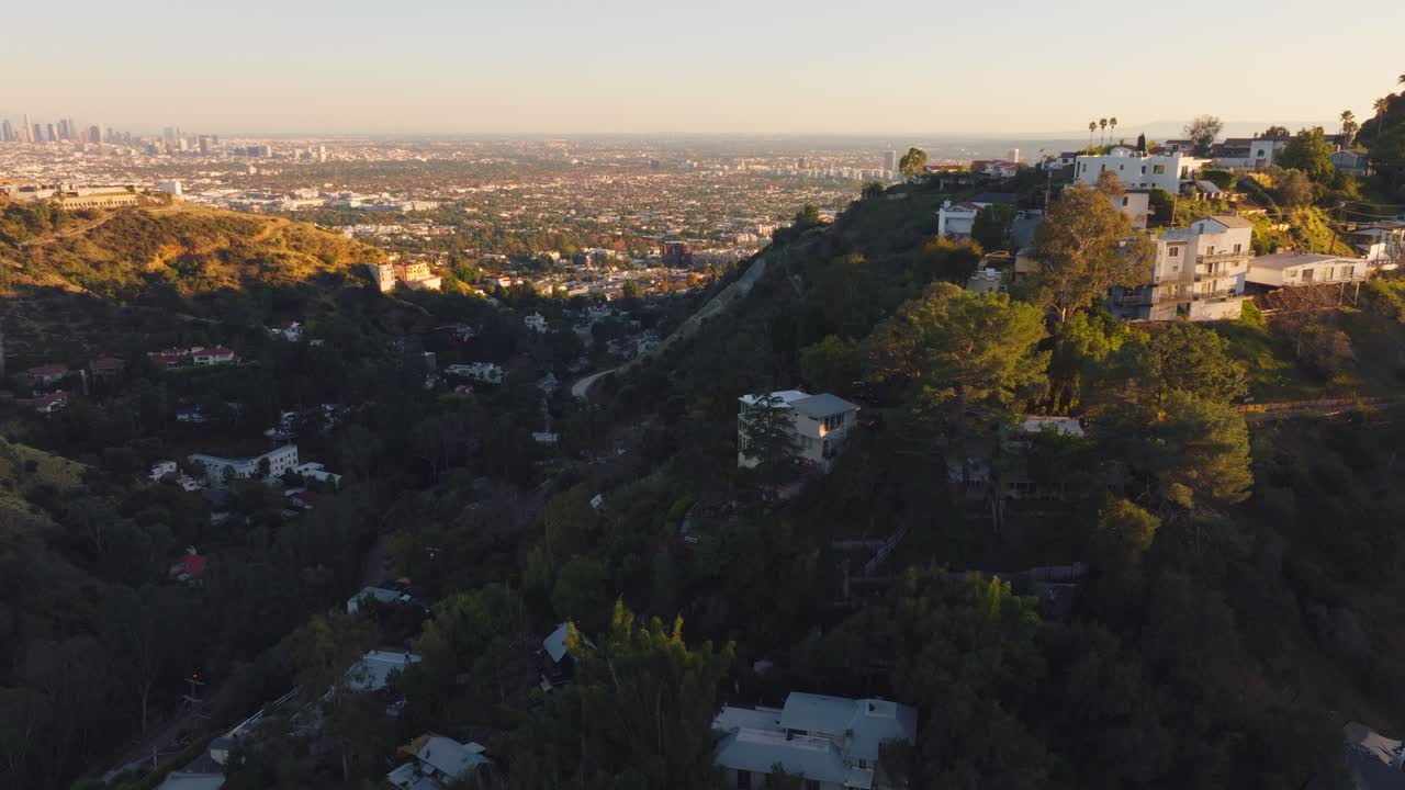 Volo sopra le Hollywood Hills all'ora d'oro, tenute di lusso immerse in colline boscose con un tramonto dorato all'orizzonte