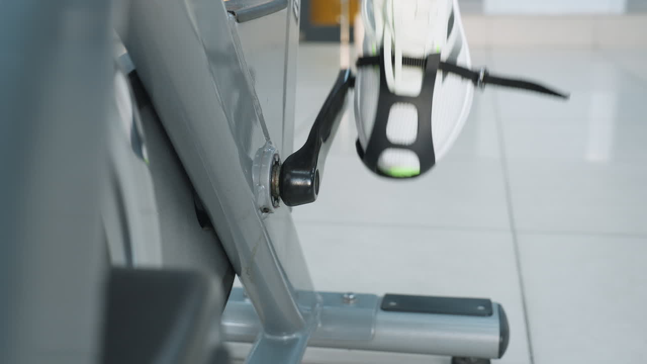 leg view of person wearing sneaker securely strapped to pedal while cycling vigorously on stationary bike in well lit modern gym with tile floor and blurred cardio equipment in background