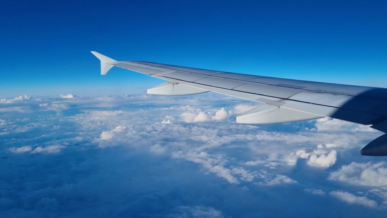 Ala di un aereo vista dal finestrino del passeggero durante il volo sopra un mare di soffici nuvole bianche sotto un cielo azzurro brillante, che evoca un senso di libertà, avventura e tranquillo viaggio aereo