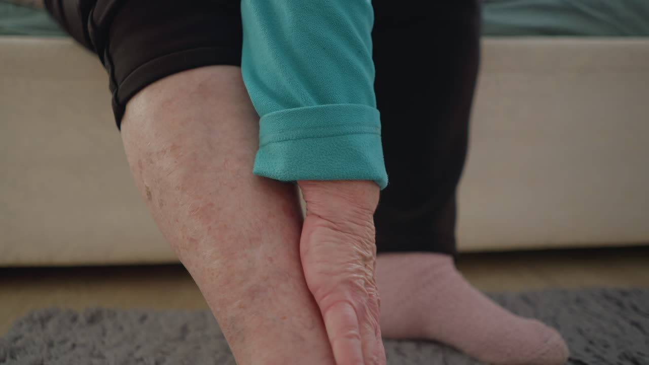 Old woman in green gently applies cream to her leg, focused on skin care while sitting on a bed. Soft light illuminates the room, creating a peaceful and comforting atmosphere for self-care