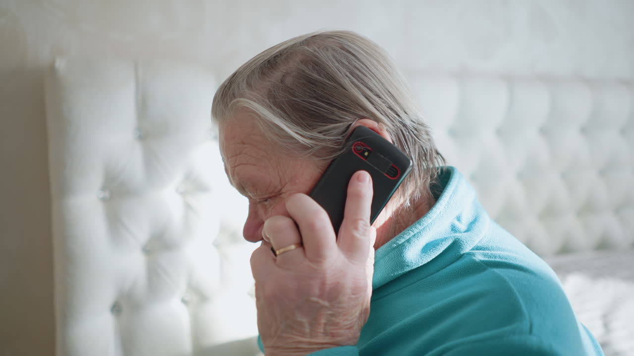 Elderly woman in green jacket talking on mobile phone, holding phone to her ear, focused expression, sitting on bed, bright and airy bedroom background, with sunlight streaming in from window