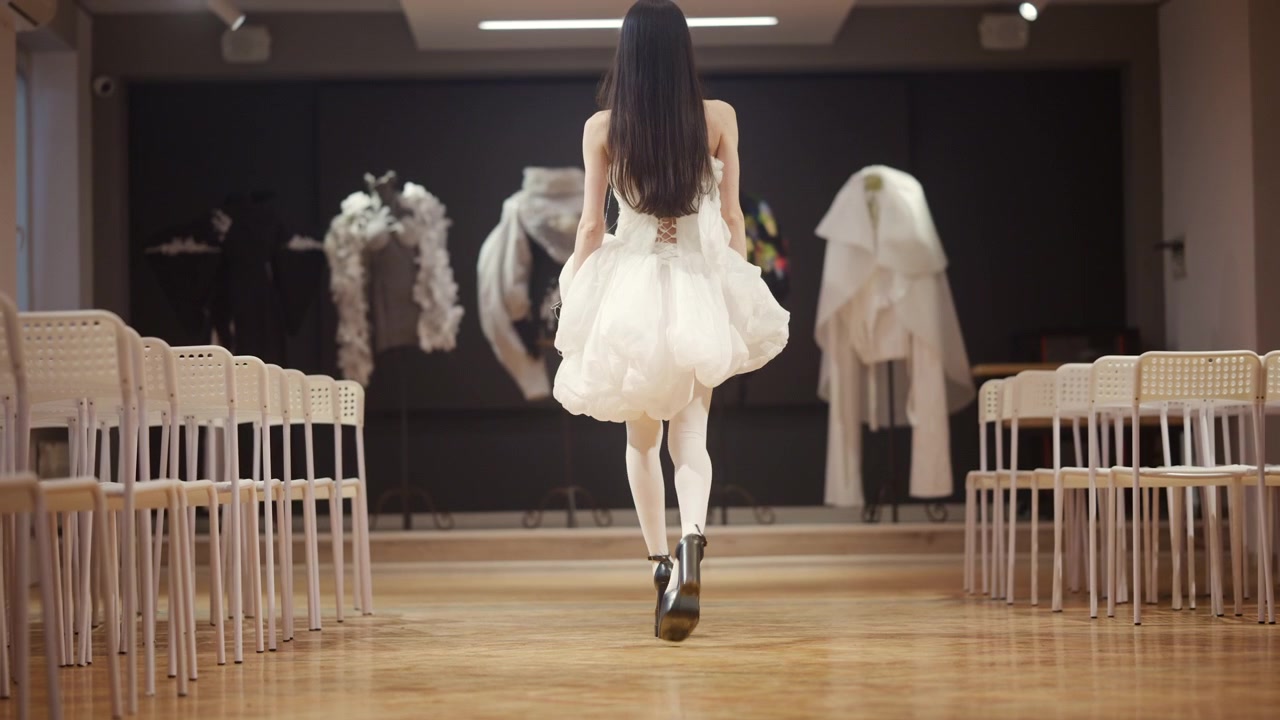 Primo piano di una modella che cammina su una passerella di legno, indossando un voluminoso abito bianco con tessuto a strati e tacchi alti neri.