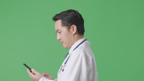 병원 에서 녹색 스크린 배경 에 걸어 다니는 동안 스마트폰 을 사용 하는 스테토스코프 를 들고 있는 아시아 남성 의사