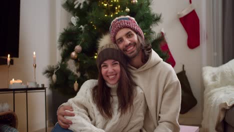 Portrait-of-a-happy-couple,-a-guy-and-a-girl-in-warm-New-Year's-hats-and-white-sweaters-are-sitting-next-to-each-other,-smiling-and-looking-at-the-camera-near-the-New-Year's-tree-in-a-cozy-apartment-in-winter