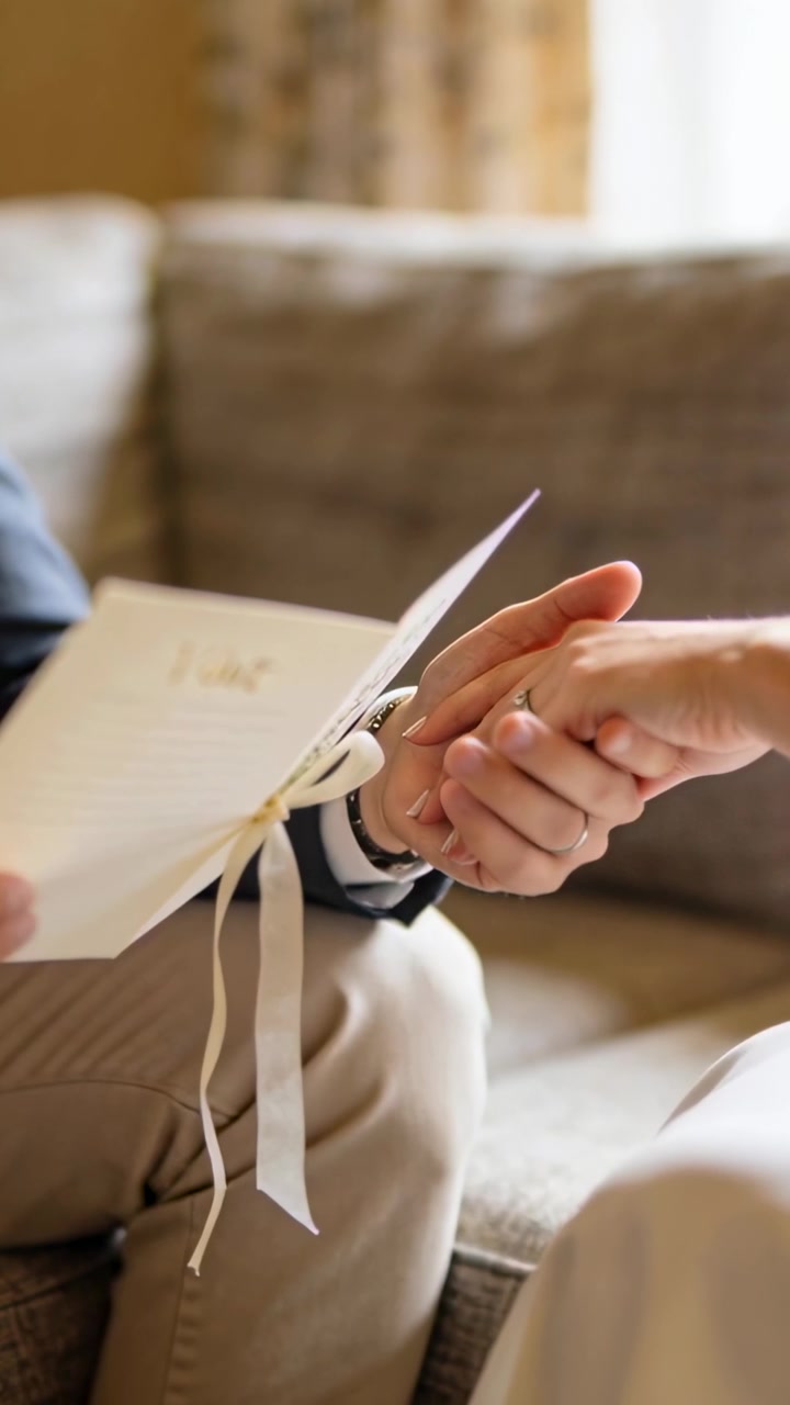 Uomo che apre un libretto legato con un nastro e accarezza la mano della donna, presentando i voti nuziali sul divano