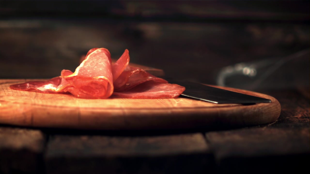 Fette di prosciutto gourmet che cadono su un tagliere di legno con un coltello.