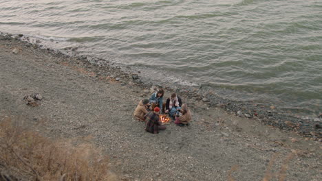 Top-View-Of-Group-Of-Teenage-Friends-Sitting-Around-The-Bonfire-While-Drinking-Tea-And-Roasting-Sausages-On-The-Seashore