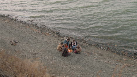 top view of group of teenage friends sitting around the bonfire while drinking tea and roasting sausages on the seashore