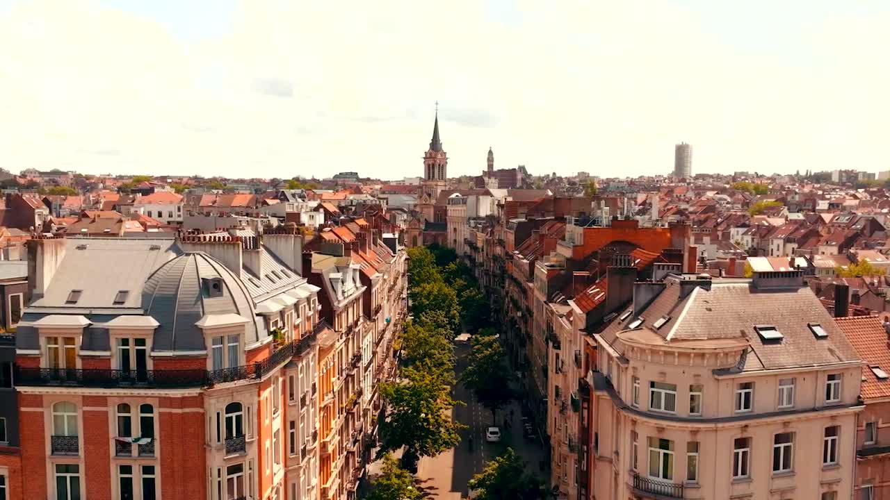 Gorgeous aerial drone footage flying in between old historic Belgium Brussels medieval houses and apartments over a neighbourhood street where green trees are towars a large church in the background.