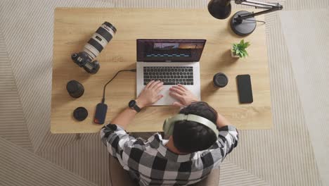 top view of a male editor with headphone shaking his head and having a headache while sitting in the workspace using a laptop next to the camera editing the video at home