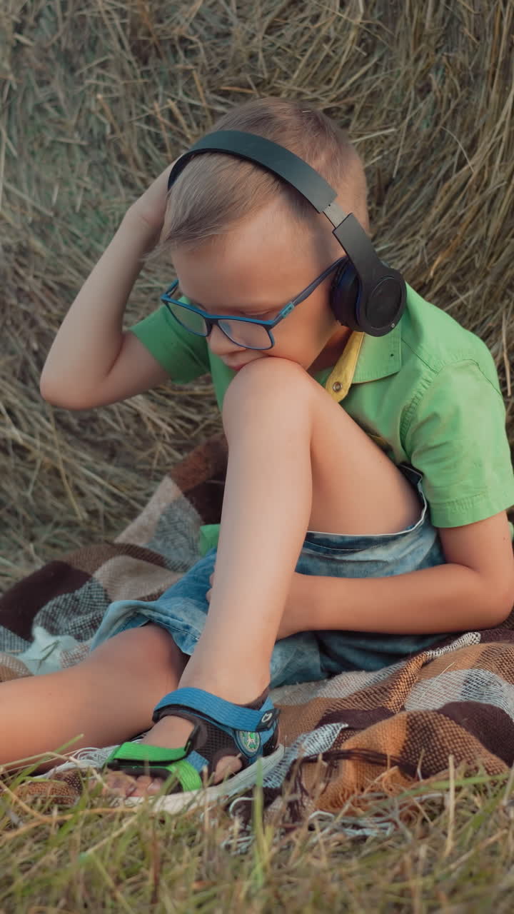 niño con auriculares y gafas, sentado en una manta, escuchando música, concentrado, con la cabeza apoyada en la rodilla y una vasta tierra de cultivo en el fondo