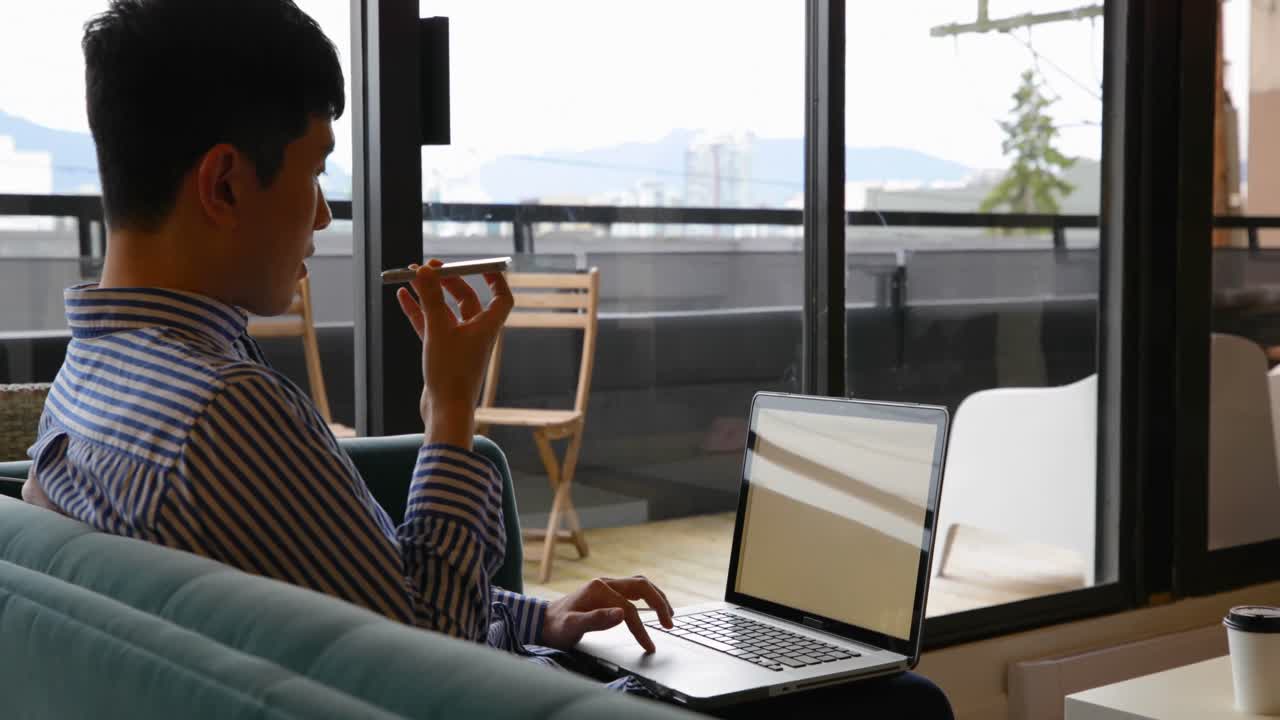 Side view of young Asian businessman talking on mobile phone and working on laptop in modern office
