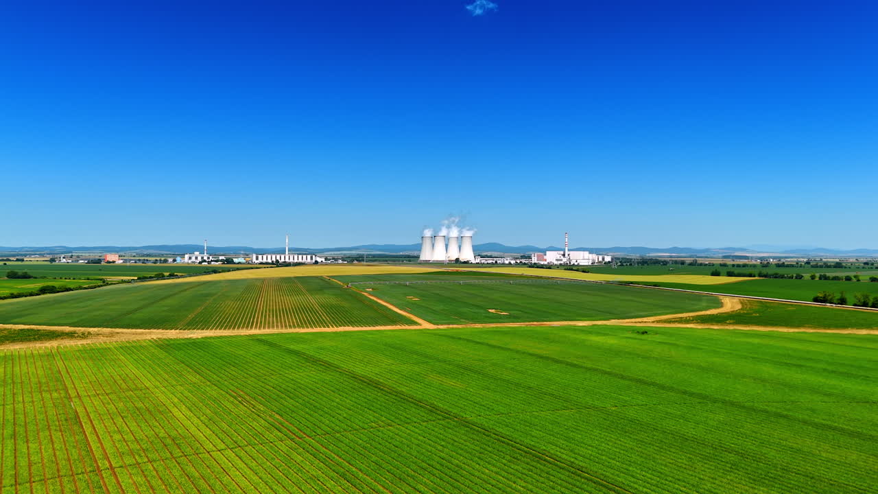 Beautiful green fields in the countryside. White smoke comes out of cooling towers of the plant at backdrop.
