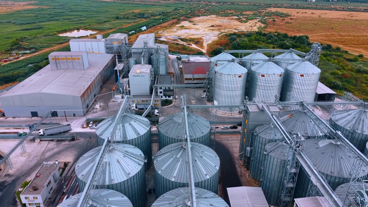 Vista aérea elevada de uma planta de elevador de grãos e silos. Múltiplos silos metálicos se erguem ao lado de edifícios de processamento industrial, próximos a campos secos.