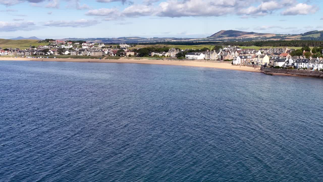 Drone pans across harbor with moored boats, waterfront village, sandy beach, and rolling hills