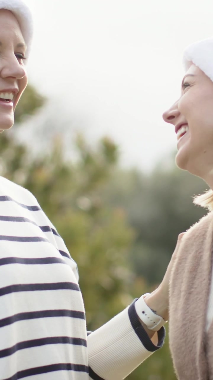 Video verticale: Due donne sorridenti, una si avvicina e tocca le spalle dell'altra, in un parco in atmosfera festiva con cappelli da Babbo Natale.