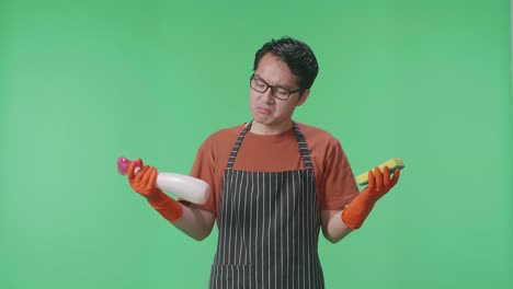 ama de llaves masculina asiática con un delantal tirando un spray y una esponja de cocina luego cruzando los brazos en el estudio de pantalla verde