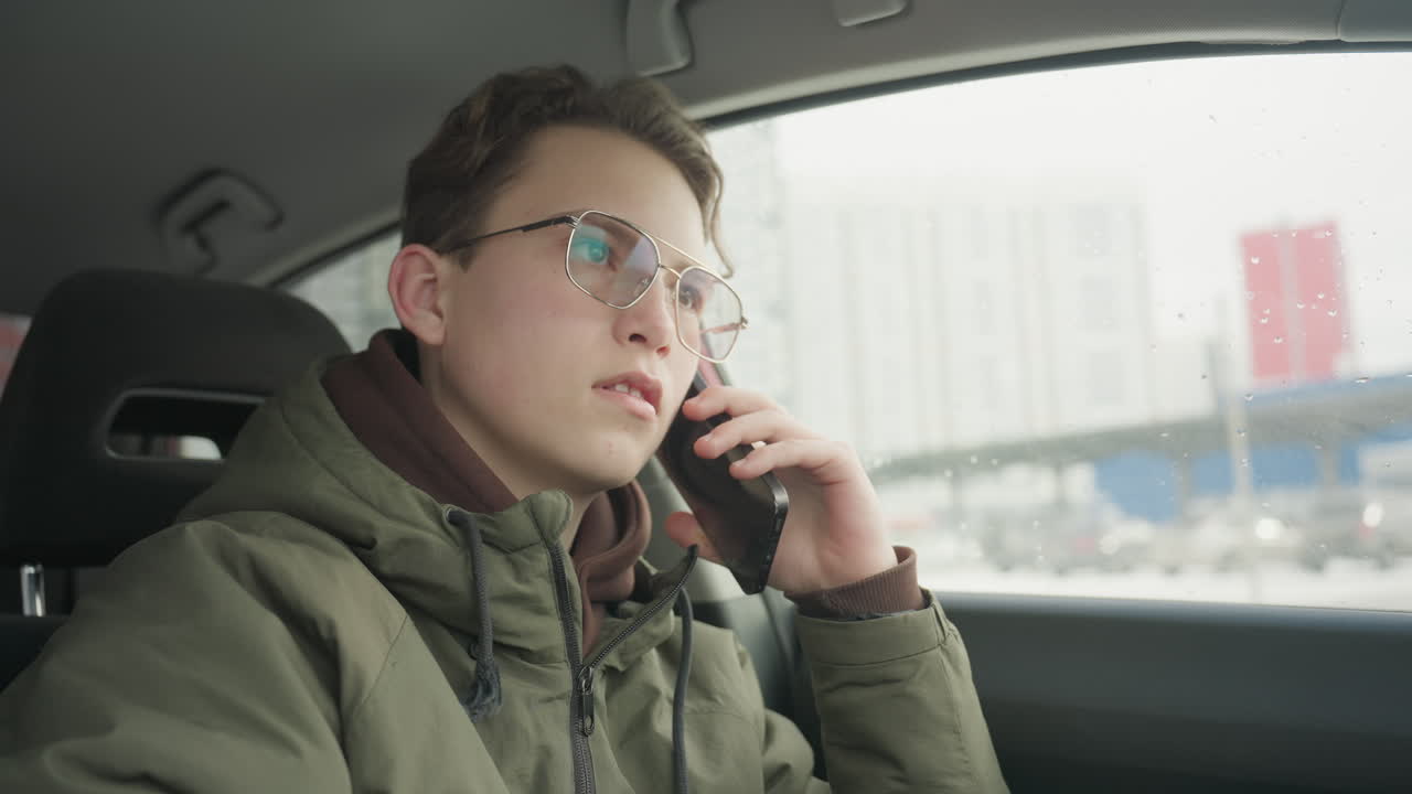 young boy in winter jacket sits inside parked car holding smartphone to ear during phone call, on cold day with cityscape and snowy background visible outside