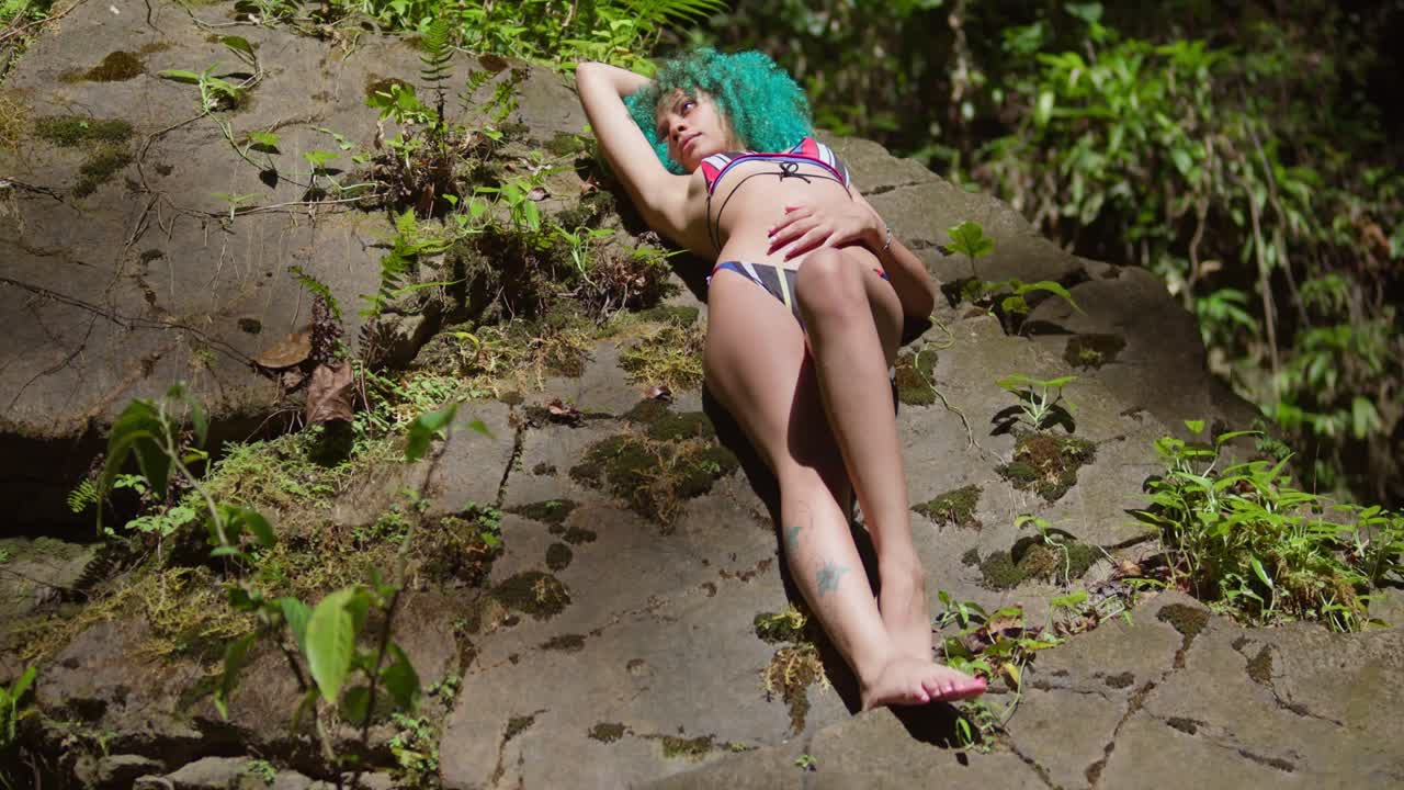 joven acostada en una roca en un bikini con el cabello rizado verde en el bosque