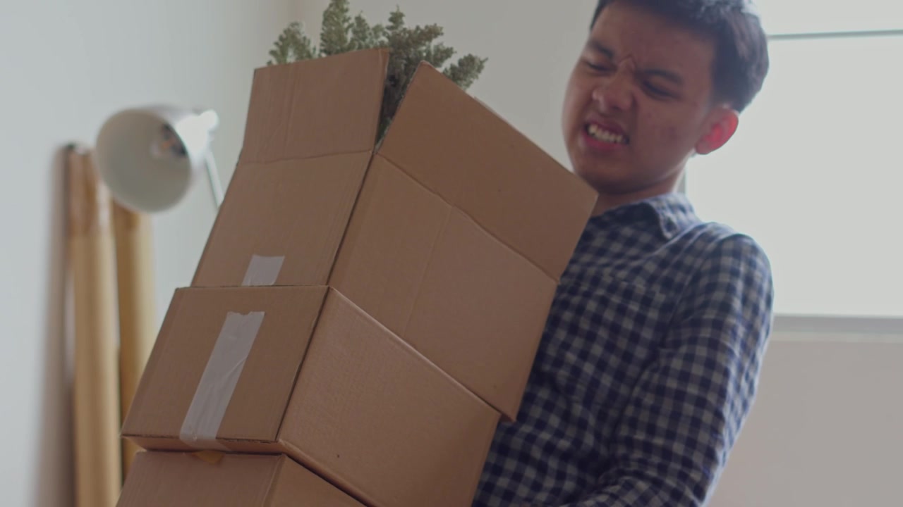 Young man struggles to lift and carry tall stack of three heavy cardboard moving boxes