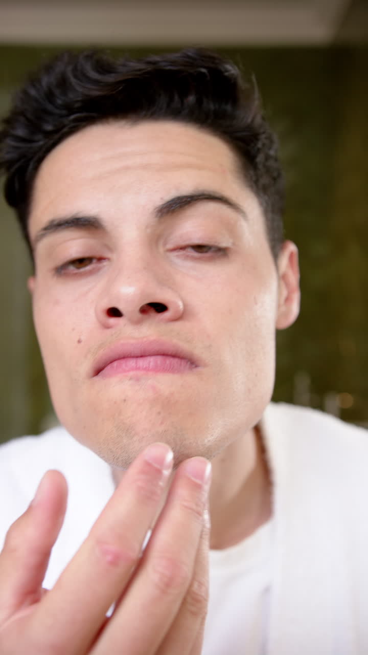 Vertical video of biracial man inspecting face and hair in bathroom mirror and smiling, slow motion