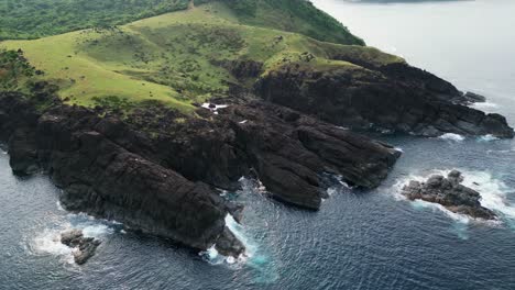 Aerial-view-of-rugged-hills-and-basalt-rocky-cliffs-facing-crashing-turquoise-ocean-waves-on-small-Philippine-island