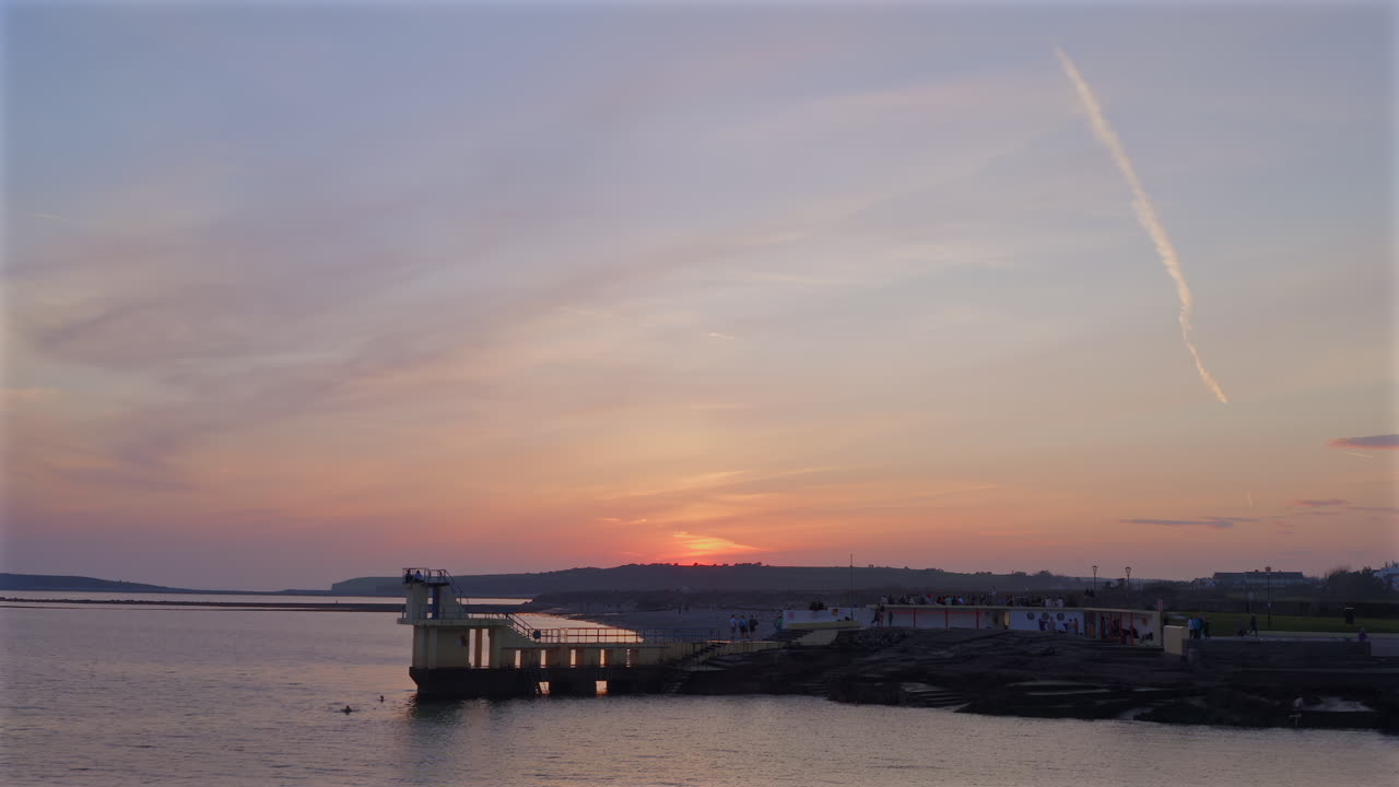 Slow motion aerial descend to Blackrock diving tower, gentle ocean waves beneath hazy sky and soft golden light, Galway Ireland