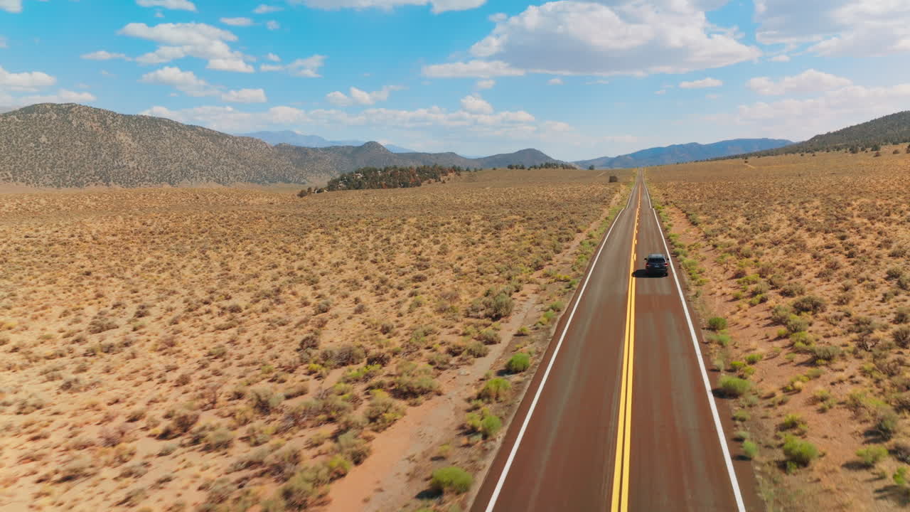 Automobile moving by the two-lane road along the dry sandy landscape. Bare mountains at backdrop of azure sky with fluffy clouds.