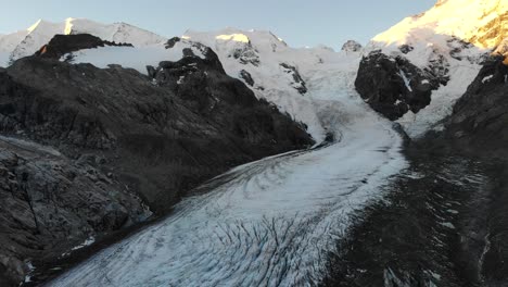 Aerial-flyover-up-away-from-the-end-of-Morteratsch-glacier-in-Engadin,-Switzerland-at-dawn-with-a-view-of-some-of-the-tallest-peaks-of-the-Swiss-Alps-like-Piz-Bernina,-Piz-Palu