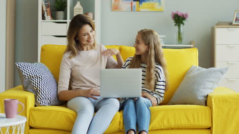 Smiled-young-woman-showing-something-to-her-teenage-daughter-on-the-laptop-computer-and-talking-with-her-while-they-sitting-in-the-living-room.-Indoors.