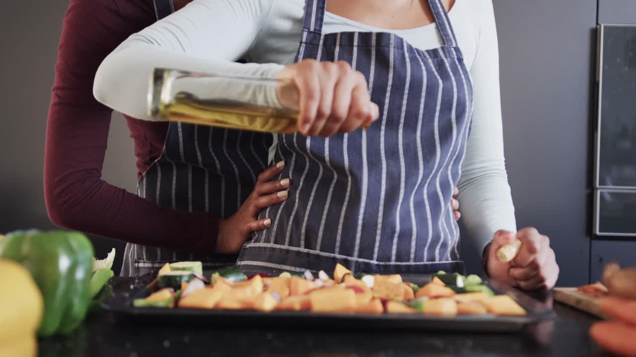 feliz pareja de lesbianas biraciales en delantales vertiendo aceite en las verduras en la cocina, en cámara lenta