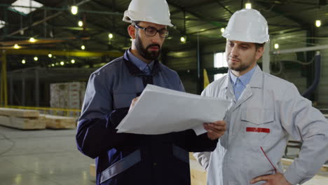 two workers wearing helmets talking while looking at blueprint in a big factory