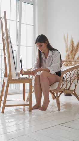 young barefoot woman art student works with paints on wooden easel sitting in comfortable rattan armchair near window in spacious studio