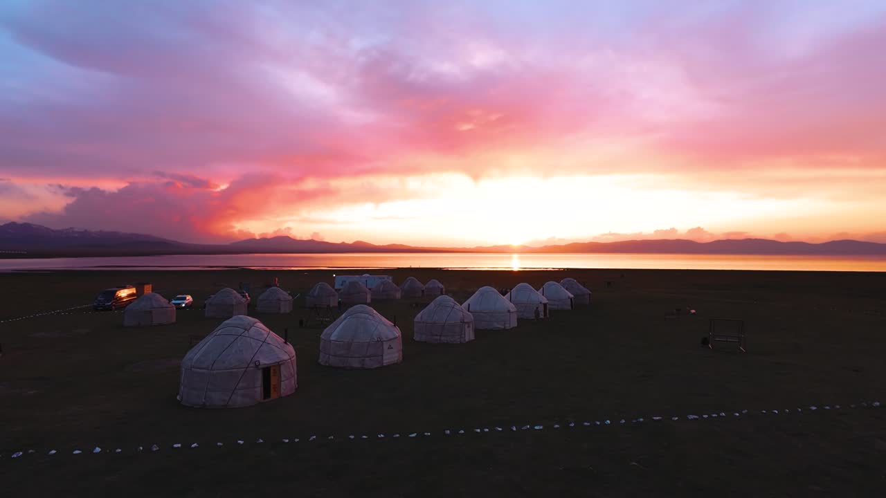 FPV drone sweeps past a yurt camp by Song Kul Lake at golden hour, warm sunset glow over the highland steppe