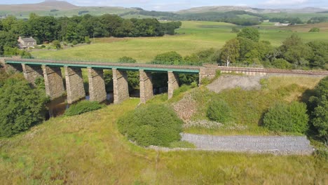 Drohnenaufnahmen-An-Einem-Sonnigen-Tag,-Die-Sich-Seitwärts-Bewegen-Und-Parallel-Zu-Einer-Ländlichen-Eisenbahnlinie-Und-Einer-Steinernen-Eisenbahnbrücke-In-Der-Landschaft-Von-Yorkshire-In-Der-Nähe-Von-Settle-Verlaufen