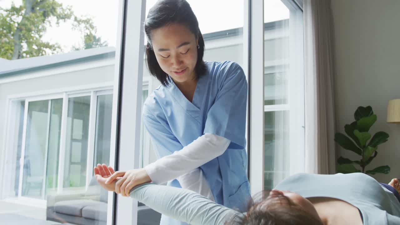 미소 짓는 아시아 여성 물리 치료사가 수술실에서 침대에 누워 있는 여성 환자의 팔을 치료하고 있습니다.