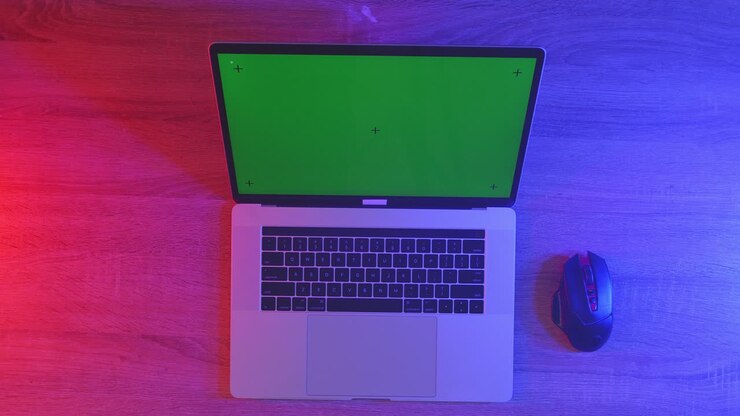 Draufsicht auf einen Laptop-Computer mit Mockup-Greenscreen-Display auf einem Holzschreibtisch, daneben ein Notizbuch mit Stiften, Brille und einem Glas Wasser. Langsames Herauszoomen, Nahaufnahme.