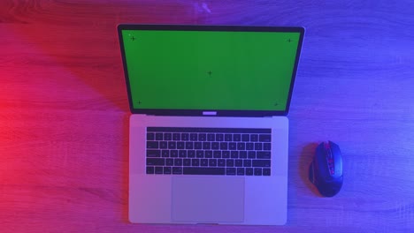 top down view of a laptop computer with mock up green screen chromakey display on a wooden office desk next to notebook with pens, glasses, and a glass of water. slow zoom out, close up