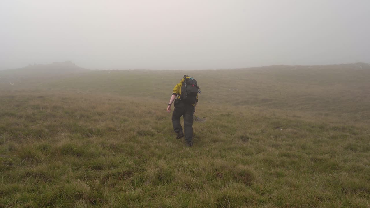 Man in Yellow Jacket Hiking up a grassy hill into the Misty