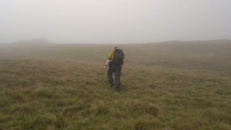 Hombre-Con-Chaqueta-Amarilla-Subiendo-Una-Colina-Cubierta-De-Hierba-Hacia-La-Niebla