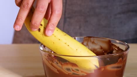 Close-up of a banana being dipped into a bowl of melted chocolate by hand.