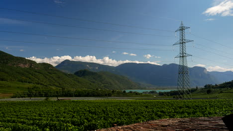 Statische-Weitwinkelaufnahme-Von-Natürlichen-Feldern,-Blauem-Himmel,-Sonnenschein-Und-Versteckten-Wolken-Hinter-Bergen-Am-Kalterer-See-In-Italien