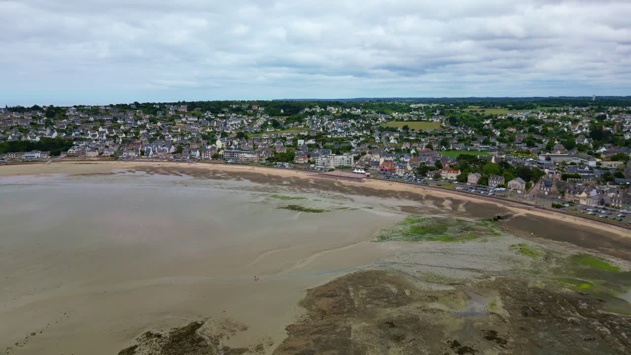Approaching drone movement to the Erquy settlement on a tidal zone beachside with coastal housing in cloudy day, low tide, Côtes-d'Armor, Brittany, France.