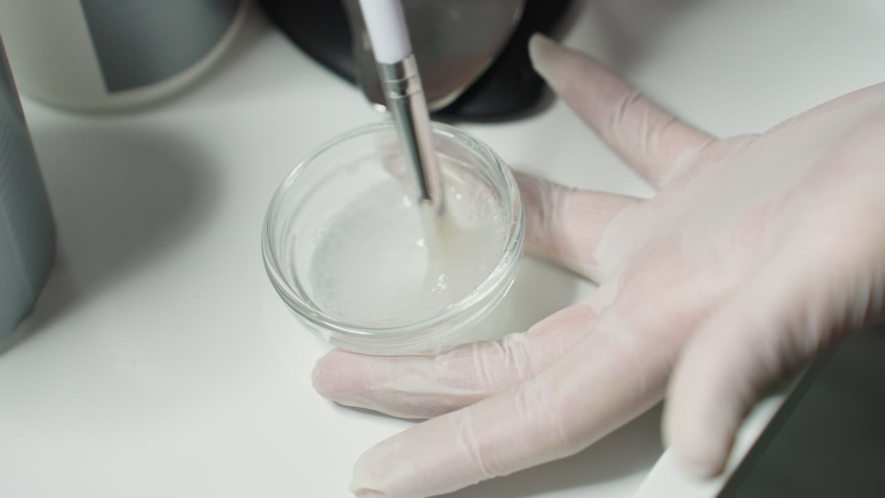 Hands on clinical gloves stir clear facial gel inside glass container on white spa tray while esthetician stirs in precise circular motions under soft light for luxurious professional care