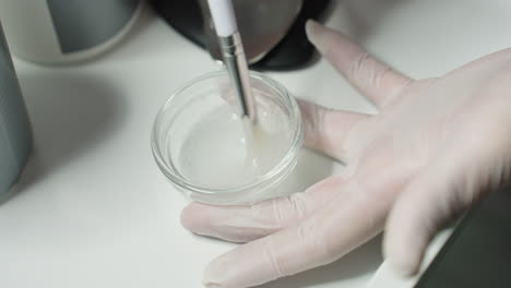 hands on clinical gloves stir clear facial gel inside glass container on white spa tray while esthetician stirs in precise circular motions under soft light for luxurious professional care