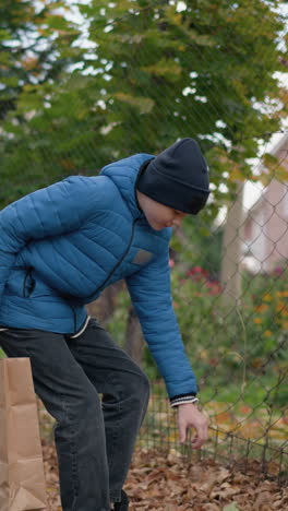 joven con chaqueta azul y gorra se inclina para recoger la hoja oxidada del suelo y la coloca en una bolsa de papel, con la valla de la barra y el edificio en el fondo, rodeado de hojas de otoño y vegetación