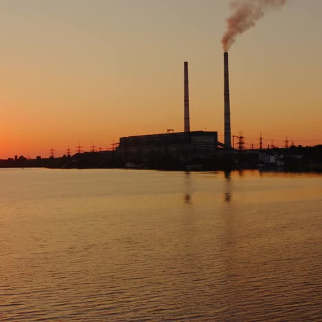 evening river on power plant background. smoke from industrial pipes goes into the air against the setting orange sun. silhouette of a factory near the lake.
