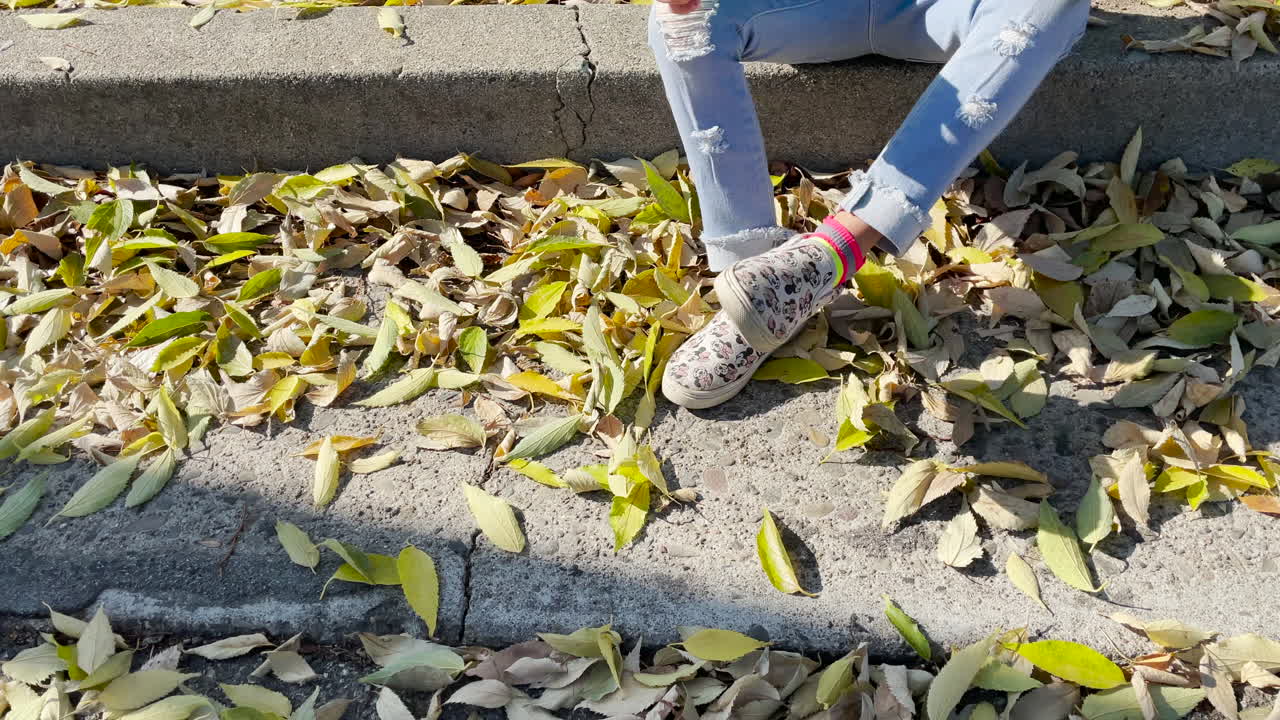 Young girl sitting on the edge of the sidewalk with leaves around her feet